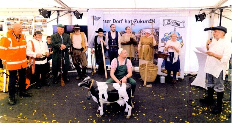 Foto von einer Schauspielgruppe, die im Kreis stehen. Im Vordergrund eine Figur einer Kuh.