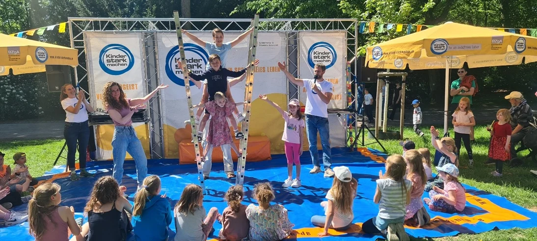 Foto von früherem Familientag. Darauf sind viele Menschen und spielende Kinder zu sehen am Stand von »Kinder stark machen«. Kinder klettern auf einer Leiter und präsentieren sich wie bei einer Show