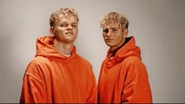Zwei junge Männer stehen in orangen Pullovern lässig da und schauen in die Kamera.
