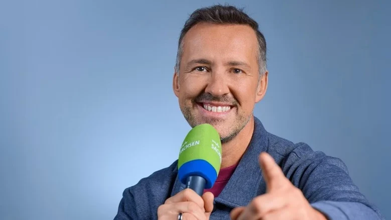 Moderator hält ein Mikrophon in der Hand und lacht in die Kamera