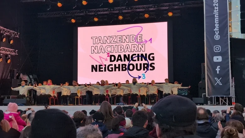 Bühnenauftritt der Gruppe „Tanzende Nachbarn / Dancing Neighbours“ vor großem Publikum mit rosafarbenem Hintergrund und Social-Media-Hinweisen.