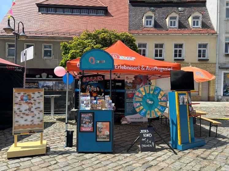 Infostand mit orangefarbenem Zelt, Drehscheibe, Plakaten und Schildern auf einem Marktplatz vor historischen Gebäuden.